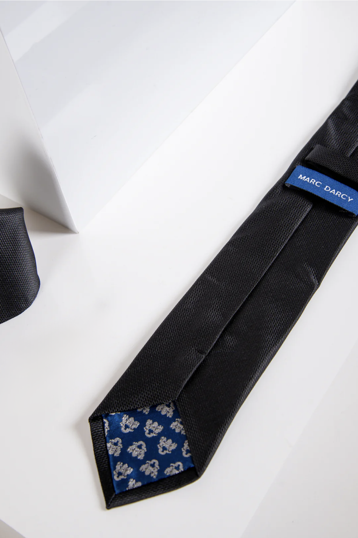 The TB17 Birdseye Tie and Pocket Square Set in Black by Accessory, featuring a blue floral inner tip, is shown on a white surface.