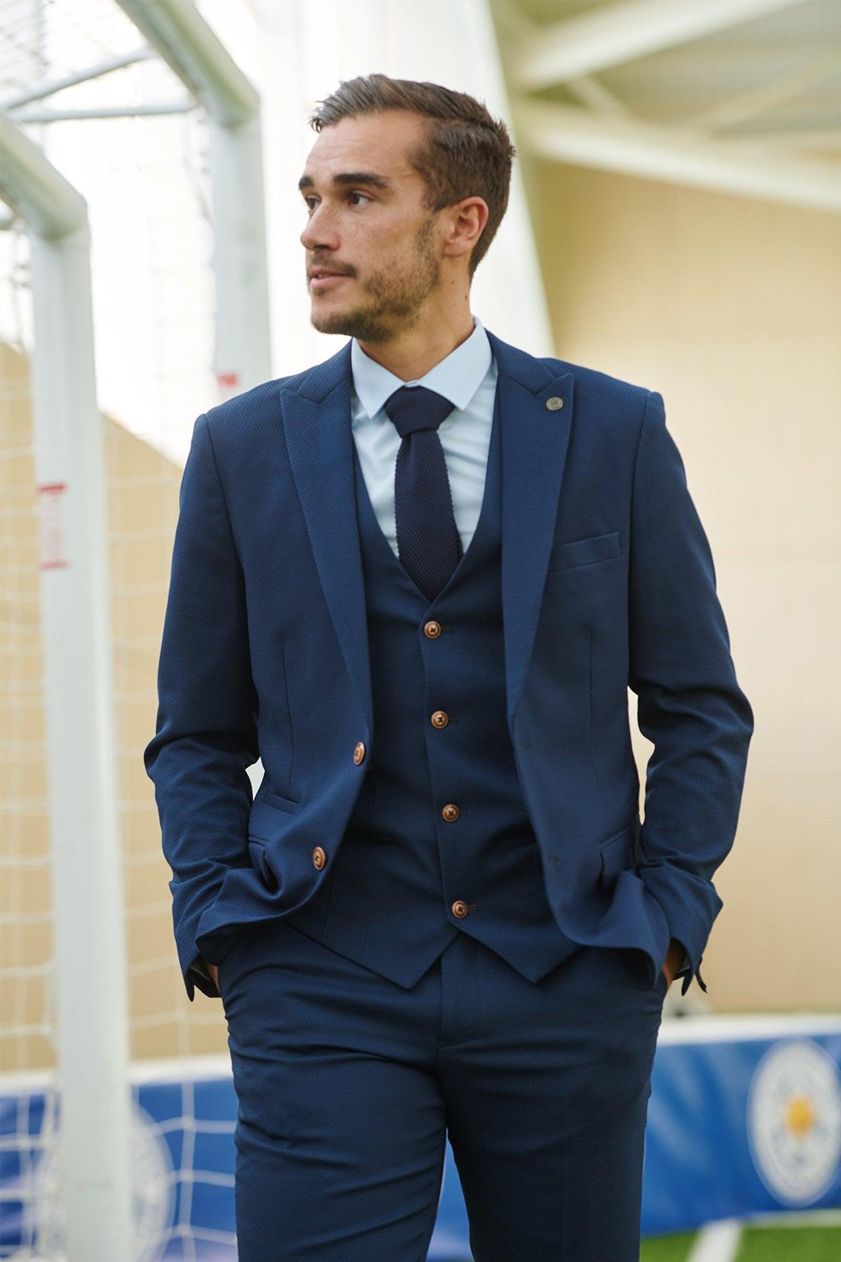 A man wearing the marcdarcy Leicester City F.C. Collection MAX Royal Blue Suit, as worn by Harry Winks, stands outdoors near a soccer goalpost with his hands in his pockets, looking to the side.