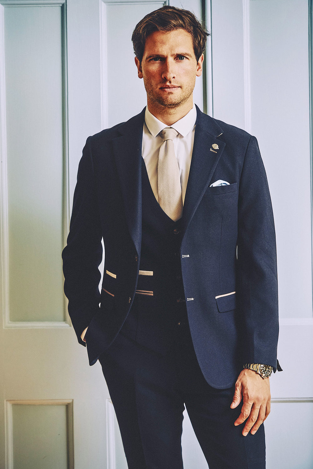 A man in the marcdarcy MD7 Navy Blue Three Piece Suit stands confidently with one hand in his pocket, wearing a white shirt and light-colored tie against a light paneled wall.