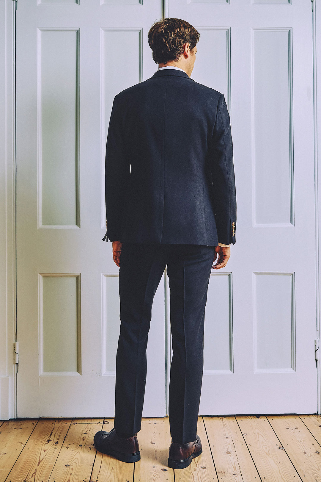 A man in a marcdarcy MD7 Navy Blue Three Piece Suit and dress shoes stands with his back to the camera in front of white paneled doors on a wooden floor.