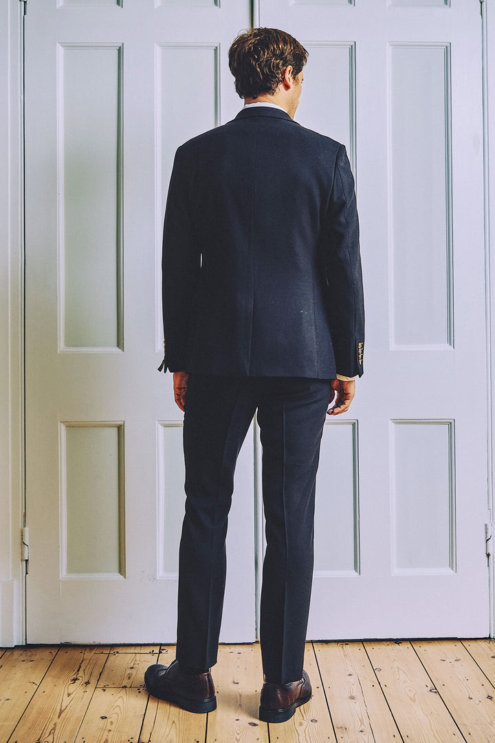 A man in a marcdarcy MD7 Navy Blue Three Piece Suit and dress shoes stands with his back to the camera in front of white paneled doors on a wooden floor.