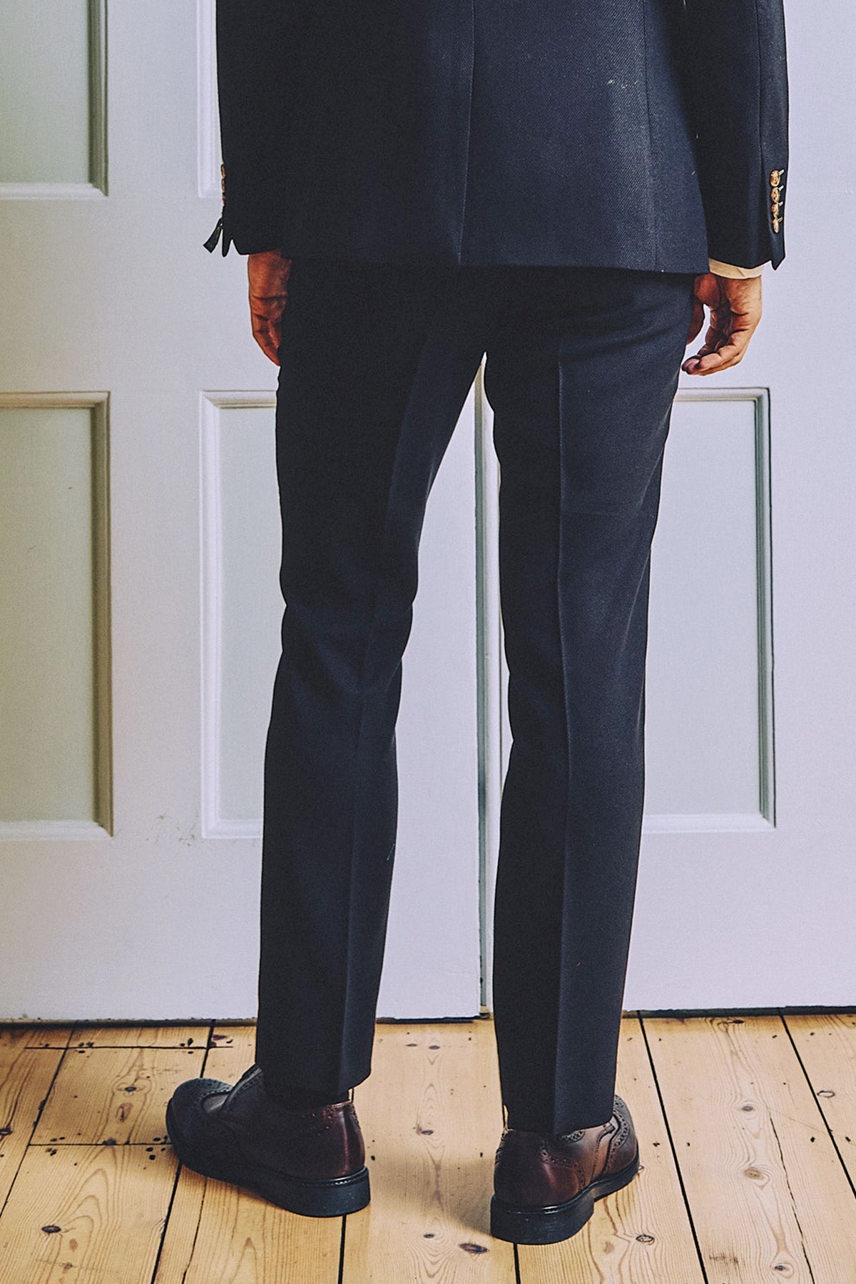 A person wearing marcdarcy MD7 - Navy Blue Trousers and brown shoes stands on a wooden floor facing a white paneled door, shown from the back.