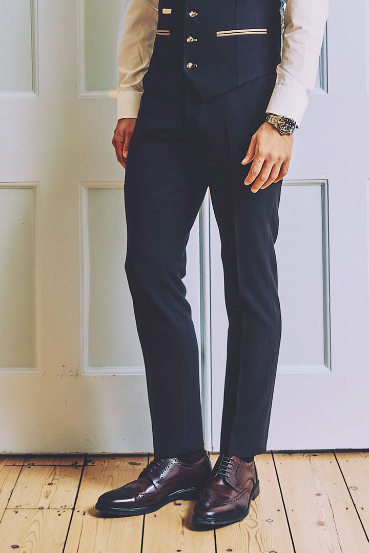 A man stands on a wooden floor before white paneled doors, dressed in formal attire featuring marcdarcy's MD7 - Navy Blue Trousers, paired with a dark vest, white shirt, brown leather shoes, and a wristwatch.