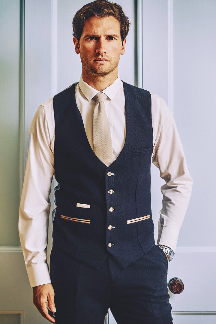 A man wearing the marcdarcy MD7 Navy Blue Three Piece Suit stands before a pale blue paneled door, pairing it with a white shirt, light tie, and dark vest featuring button and pocket details, looking directly at the camera.
