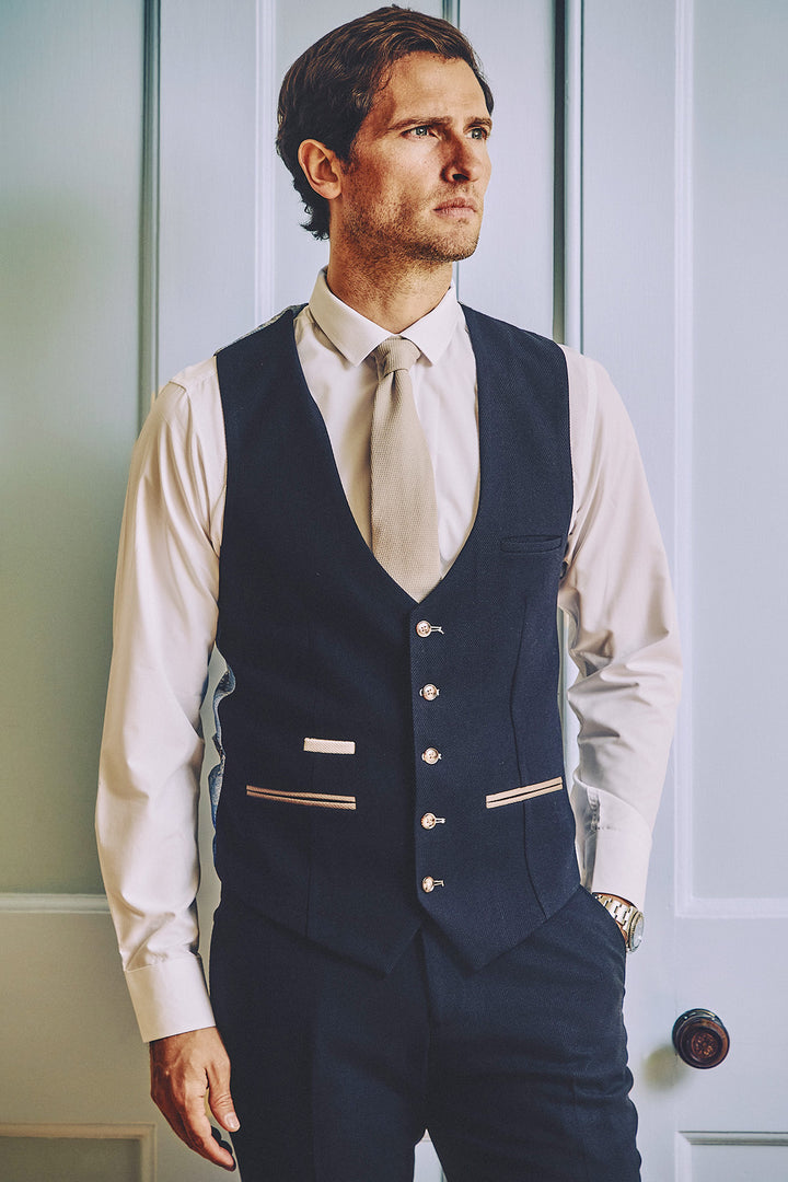 A man wearing the Marc Darcy MD7 - Navy Blue Waistcoat, paired with a white dress shirt and light-colored tie, stands in front of a light door, looking thoughtfully to the side with one hand in his pocket.