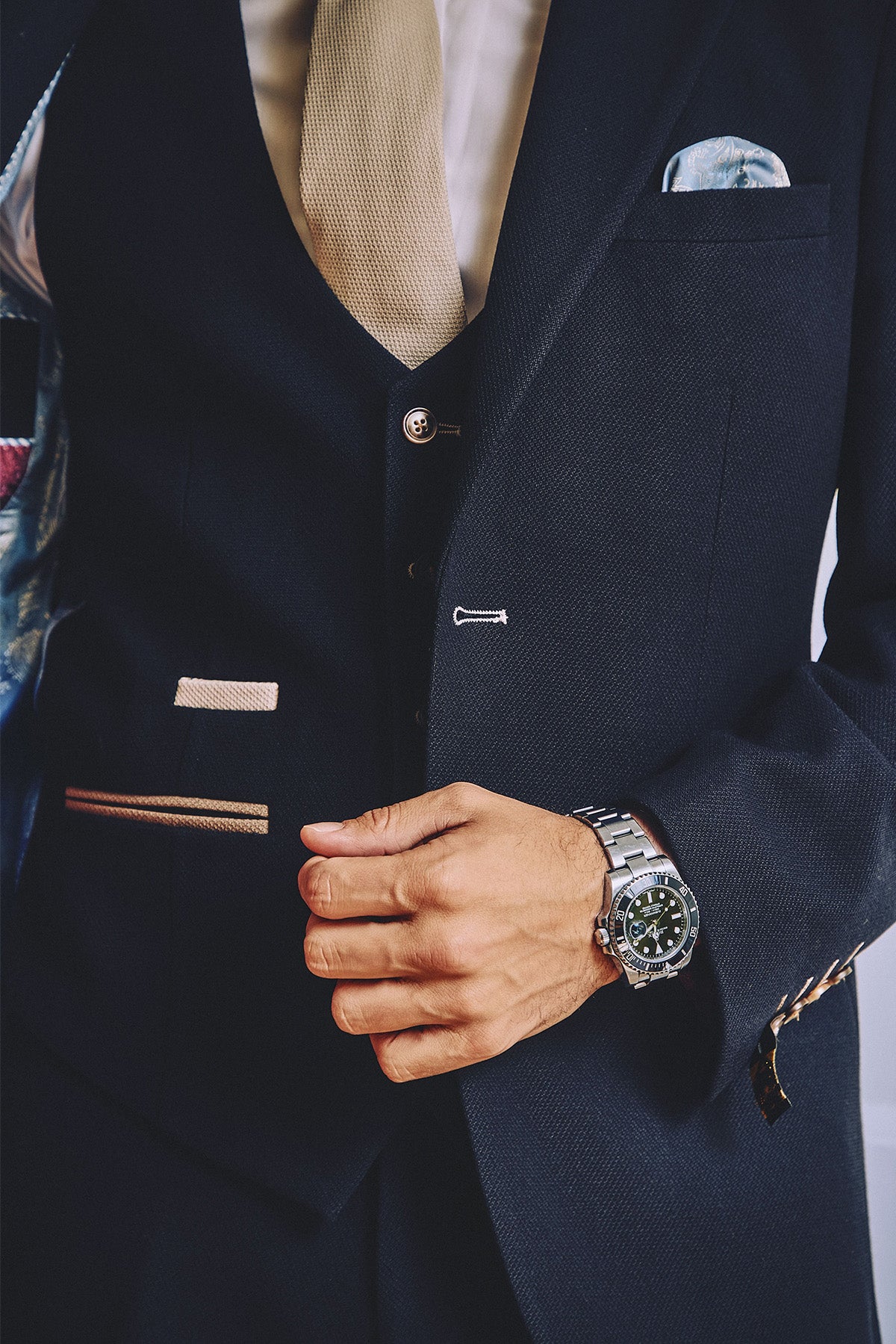 Wearing the marcdarcy MD7 Navy Blue Three Piece Suit, a person adjusts their jacket, revealing a beige tie, white shirt, silver watch, and light blue pocket square. Only their torso and hand are visible.