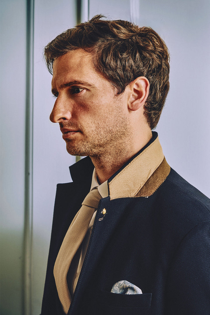 A man with short brown hair, dressed in the marcdarcy MD7 - Navy Blue Blazer featuring a contrasting tan collar, beige shirt, and tie, stands in profile against a light-colored background.