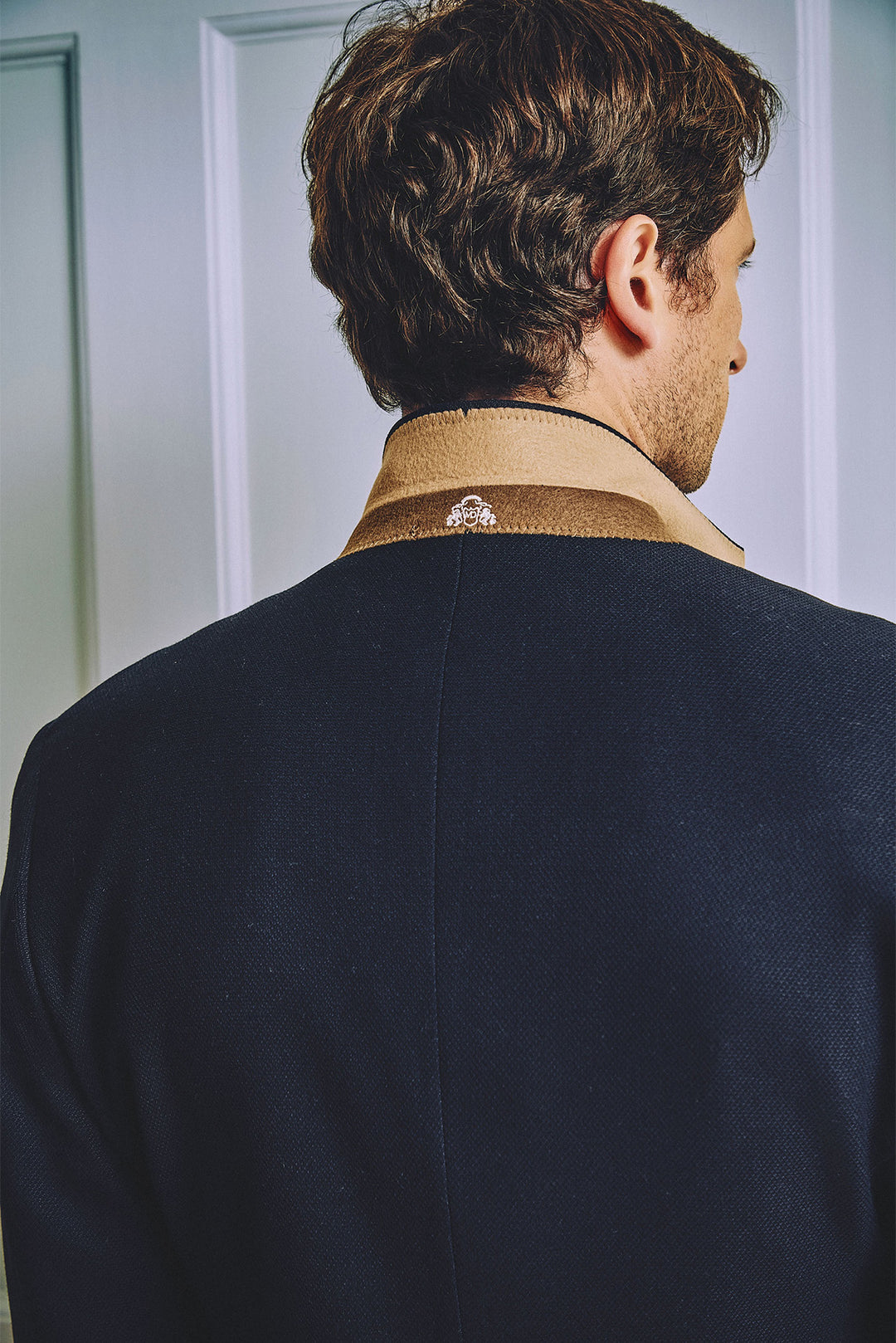 A man with brown hair, seen from behind, wears the marcdarcy MD7 Navy Blue Three Piece Suit over a tan collared shirt, standing in front of a white paneled wall.