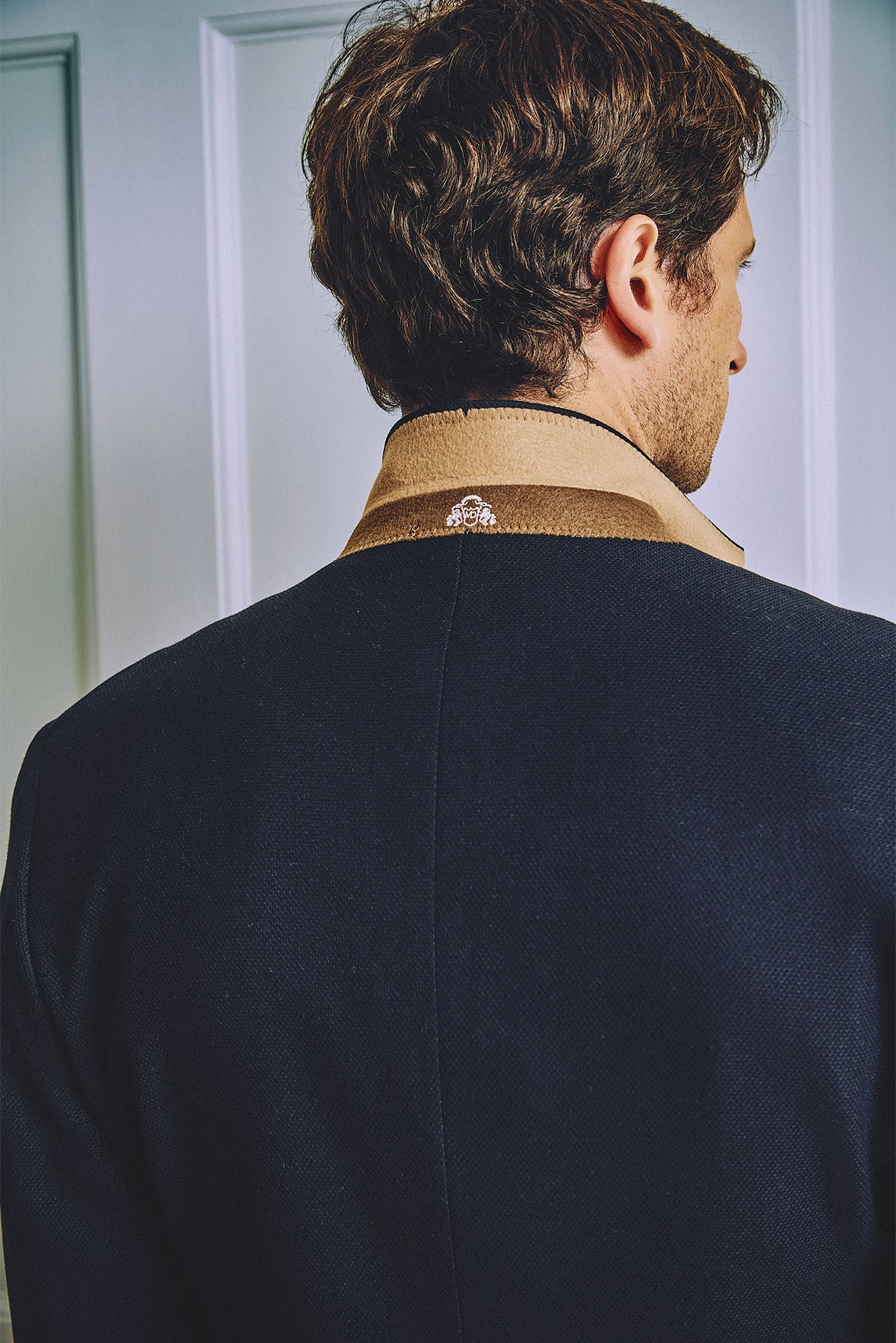 A man with brown hair, seen from behind, wears the marcdarcy MD7 Navy Blue Three Piece Suit over a tan collared shirt, standing in front of a white paneled wall.