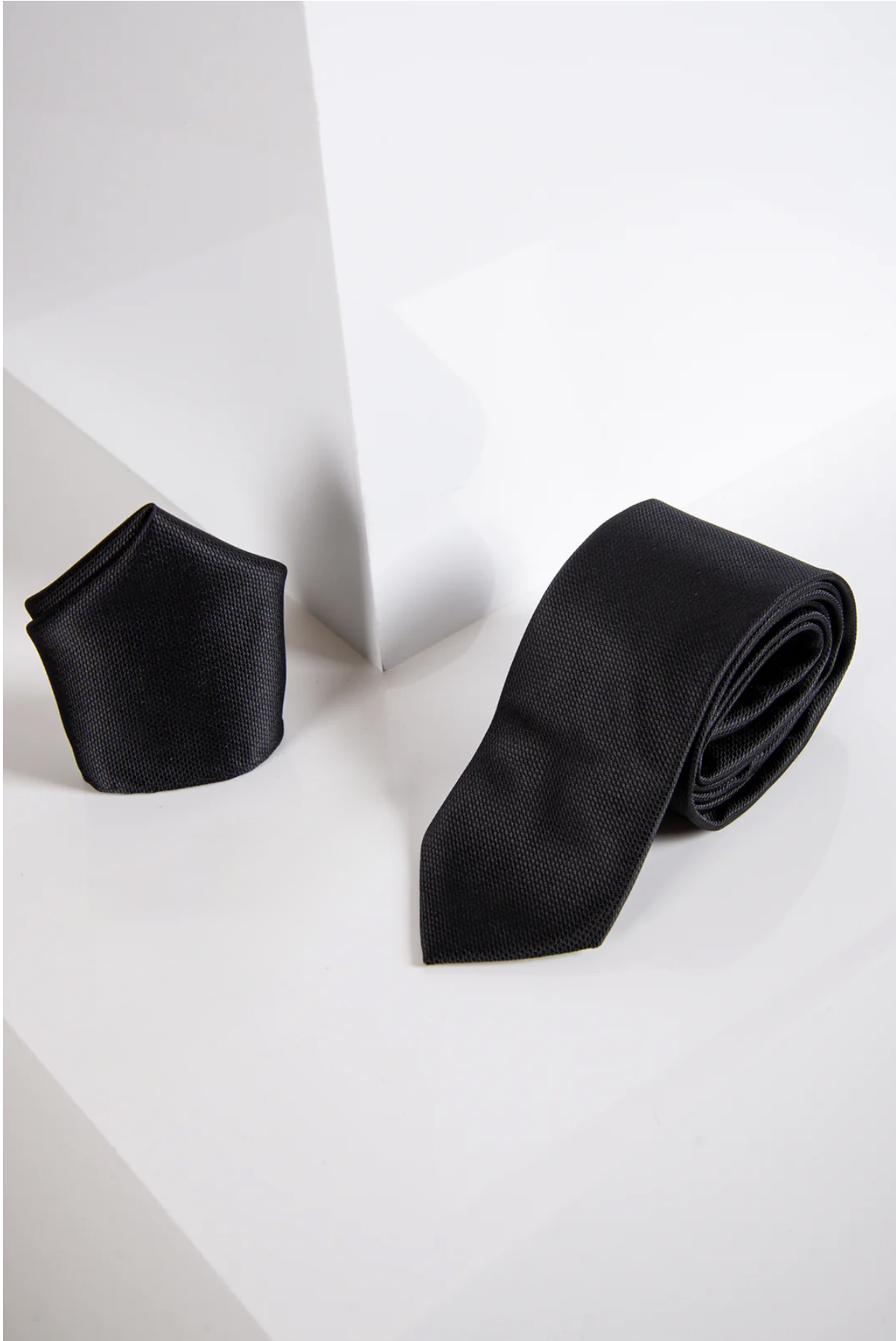 The Accessory TB17 Birdseye Tie and Pocket Square Set in Black is displayed on a white geometric surface with sharp angles and clean lines.