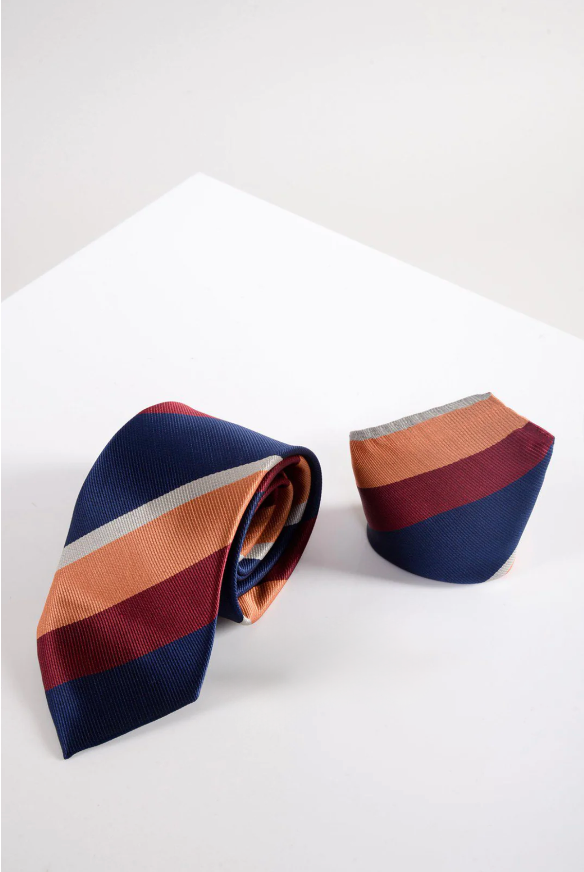 The Marc Darcy BRUNO Multicolour Stripe Print Tie and Pocket Square Set, featuring horizontal navy, orange, burgundy, and cream stripes, is shown rolled on a white surface with a geometric backdrop for a refined look.