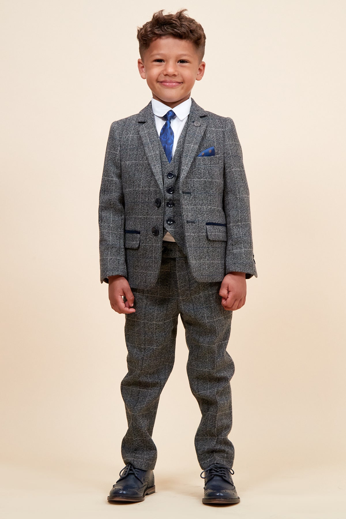 A young boy smiles in the marcdarcy SCOTT - Childrens Grey Check Three Piece Suit, paired with a white shirt, blue tie, and dark shoes, set against a plain beige background.