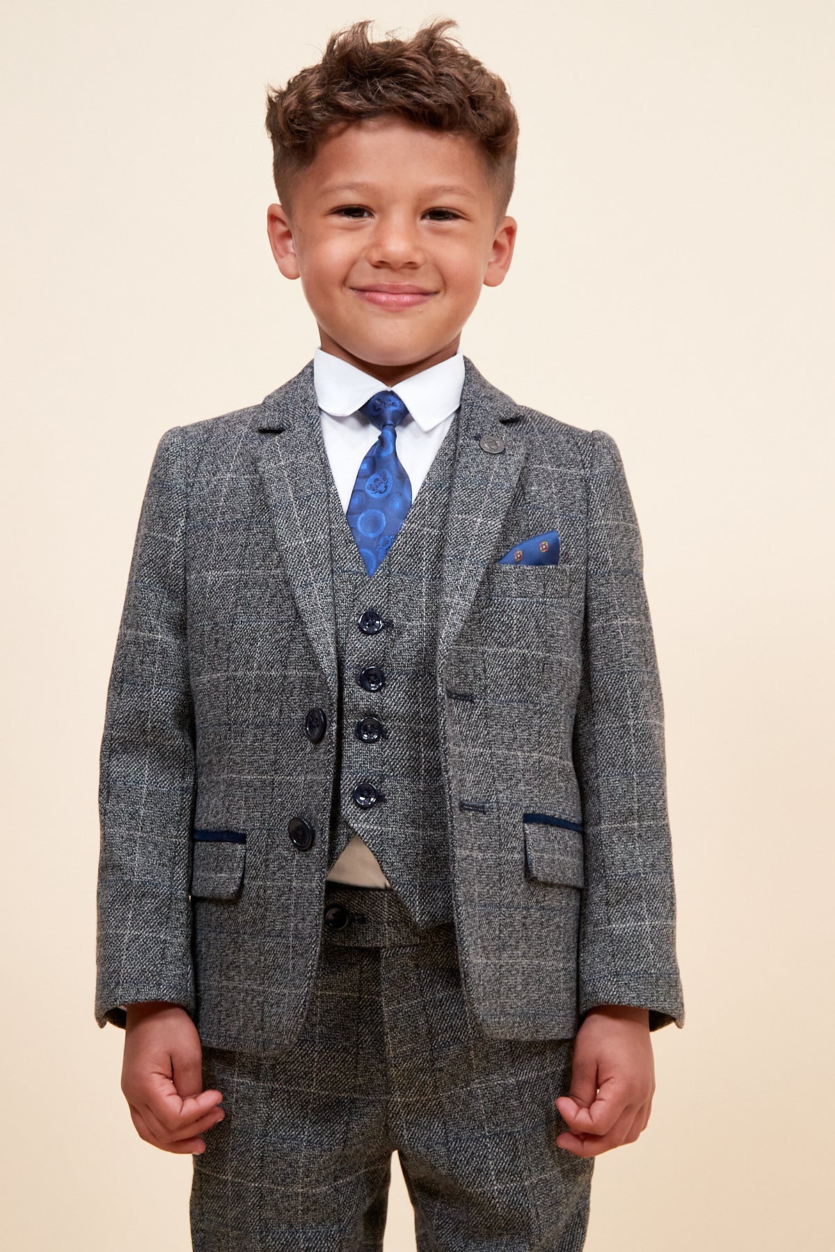 A young boy smiles in the marcdarcy SCOTT - Childrens Grey Check Three Piece Suit, featuring a matching vest, white shirt, blue patterned tie, and adjustable waistband, standing against a plain light background.