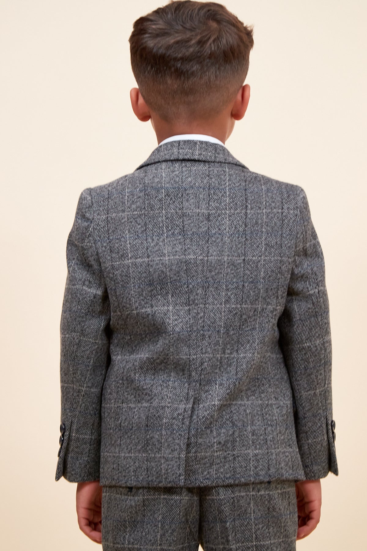 A young boy with short brown hair stands with his back to the camera, wearing the marcdarcy SCOTT Childrens Grey Check Three Piece Suit with an adjustable waistband, set against a plain light background.