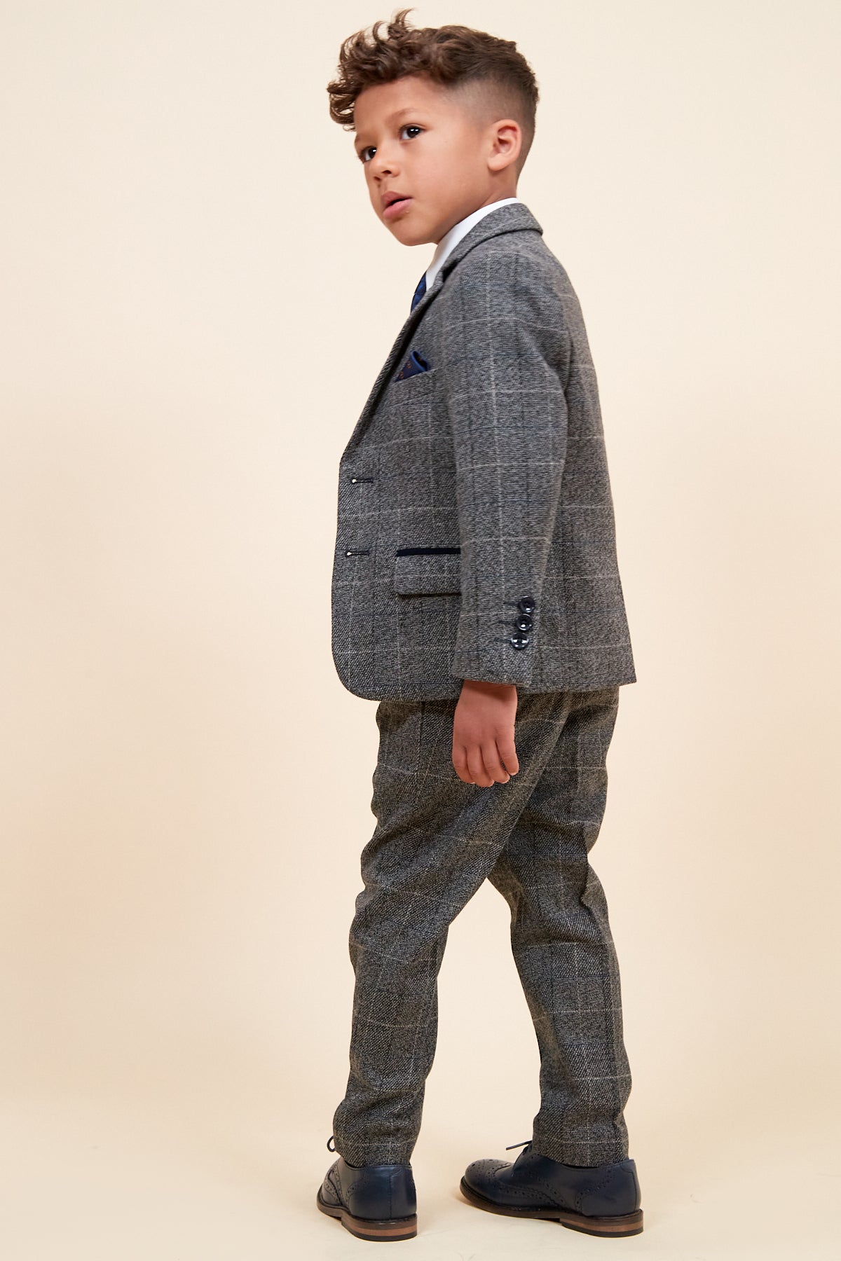 A young boy stands sideways, gazing ahead in the marcdarcy SCOTT Children’s Grey Check Three Piece Suit with matching trousers and dark dress shoes against a plain, light background.