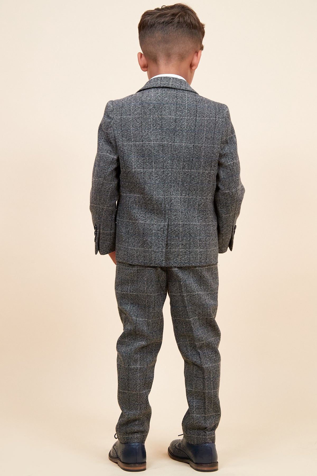 A young boy faces away from the camera, wearing the marcdarcy SCOTT Childrens Grey Check Three Piece Suit with matching pants and dark shoes, featuring an adjustable waistband, set against a plain beige background.