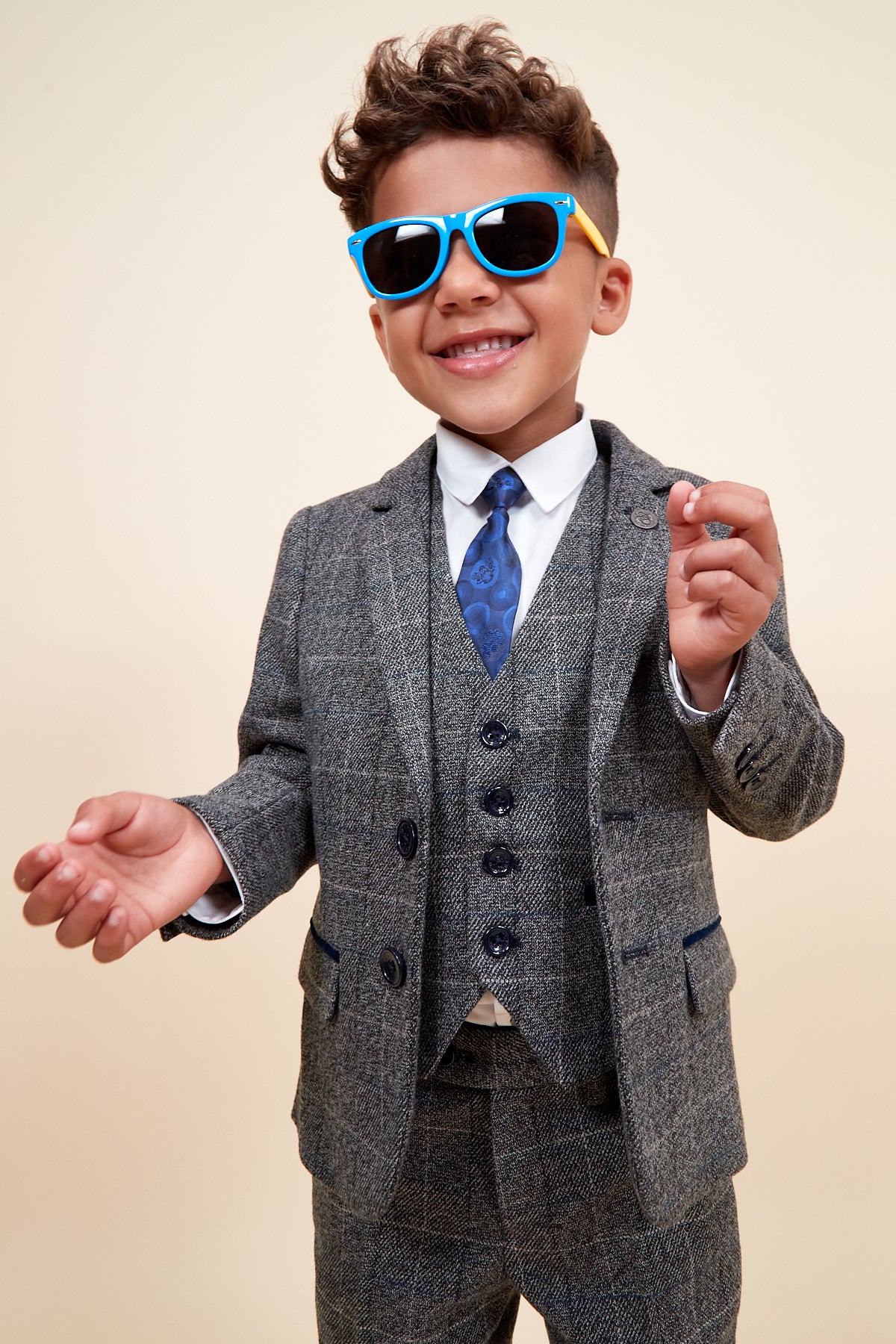 A young boy smiles wearing the marcdarcy SCOTT Children's Grey Check Three Piece Suit, paired with blue sunglasses and a patterned blue tie.