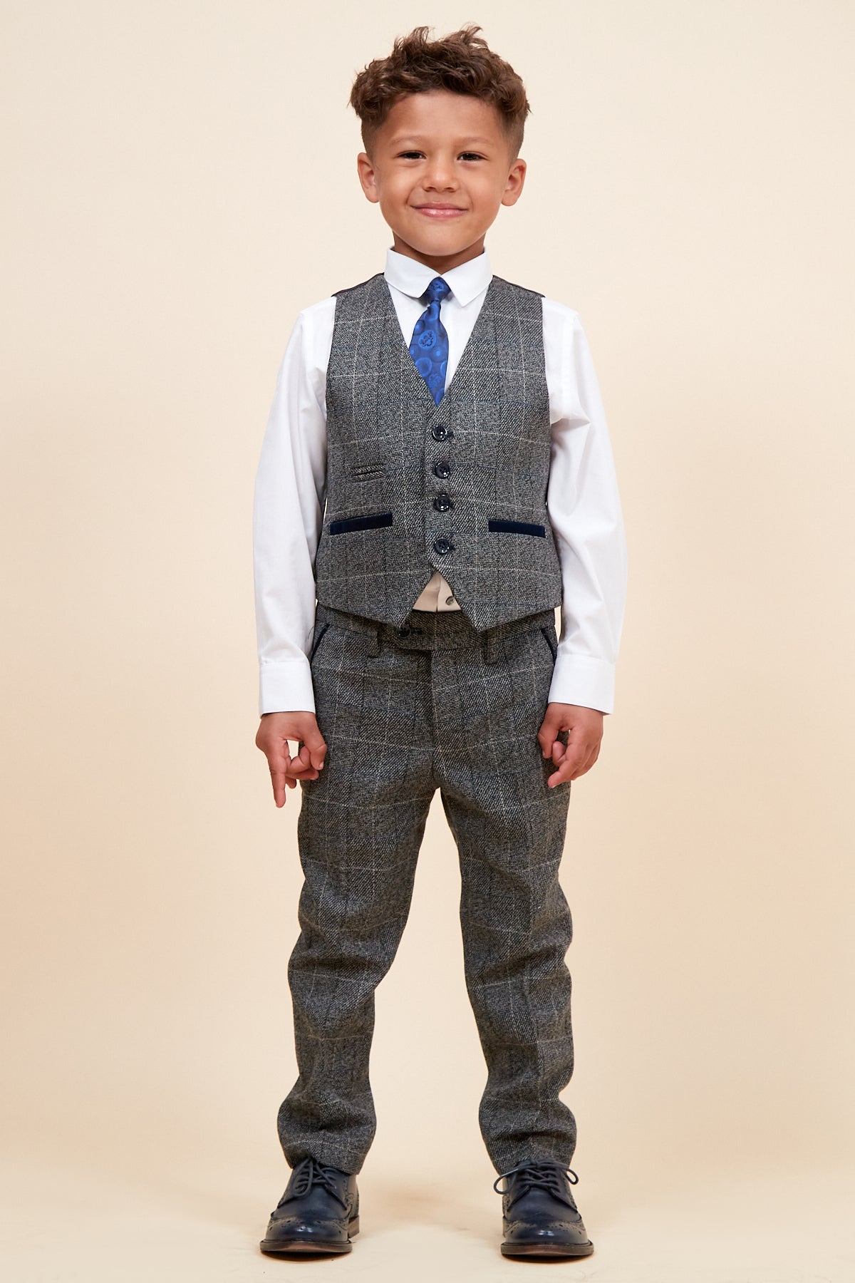 A young boy wears the marcdarcy SCOTT Children's Grey Check Three Piece Suit with adjustable waistband, white shirt, and blue tie, smiling at the camera.