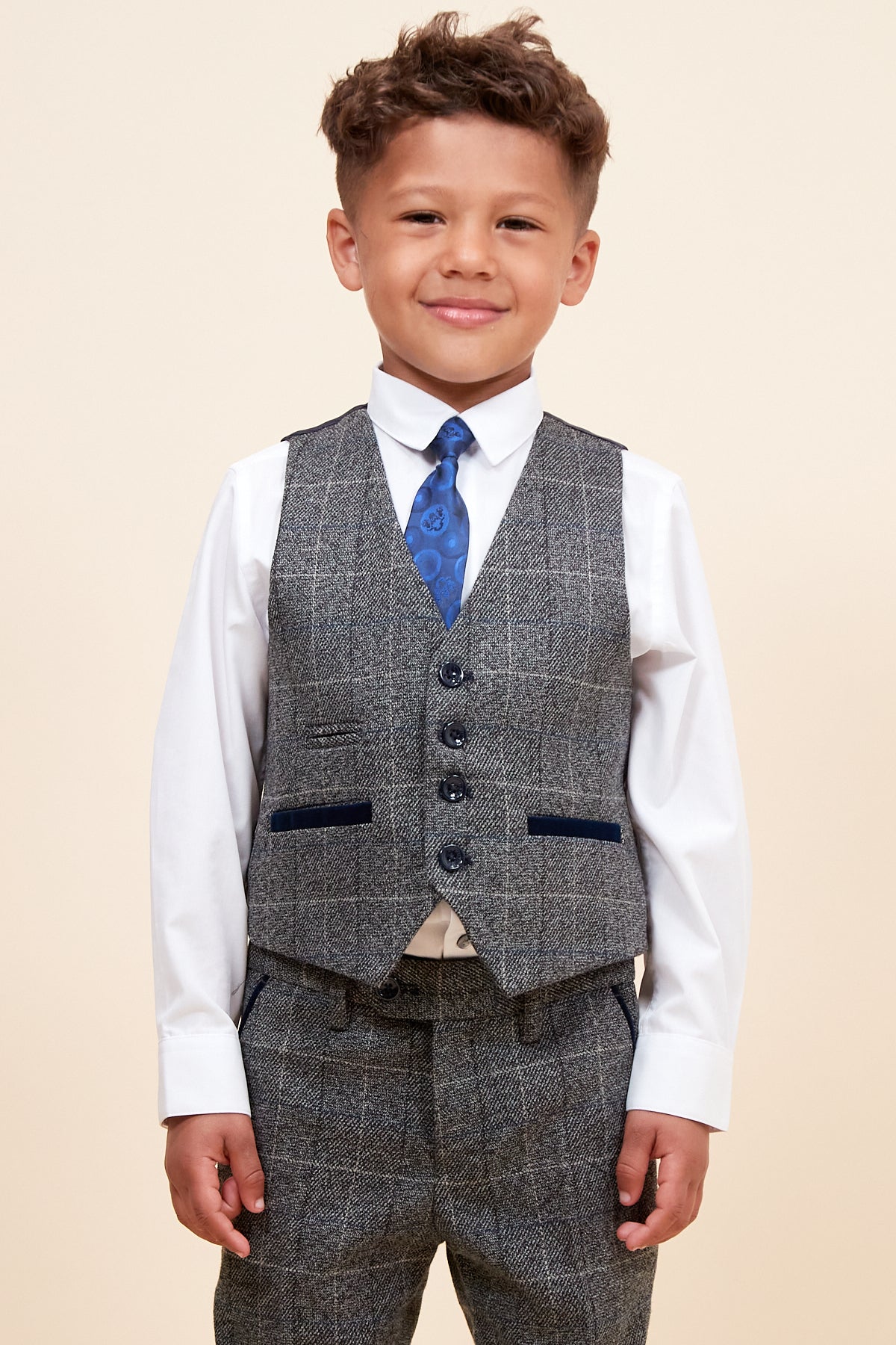 A young boy with short brown hair smiles, wearing the marcdarcy SCOTT Childrens Grey Check Three Piece Suit with an adjustable waistband, along with a white shirt and blue patterned tie, standing against a plain beige background.