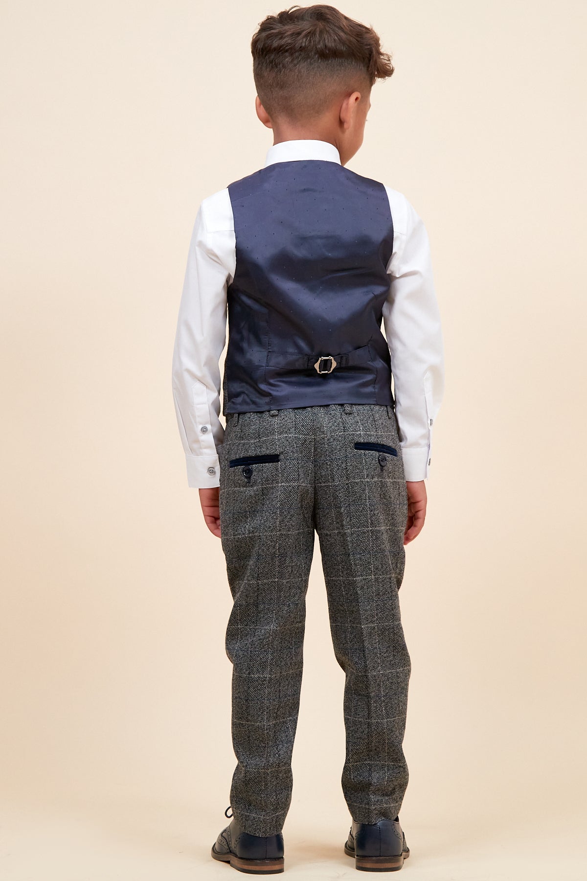 A young boy faces away from the camera, wearing the marcdarcy SCOTT Childrens Grey Check Three Piece Suit with a white shirt, gray plaid trousers, dark blue vest, and dark shoes, set against a plain beige background.
