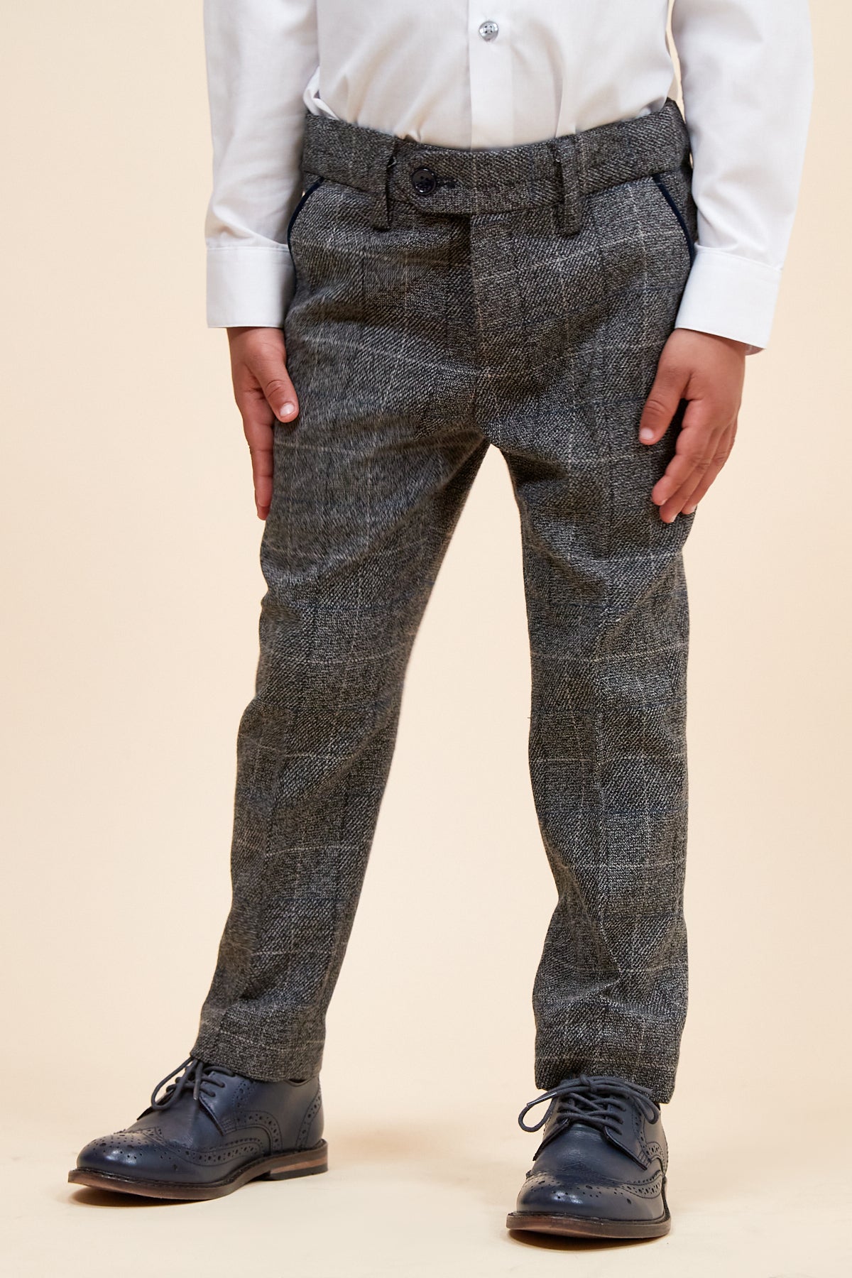 A child models the marcdarcy SCOTT - Childrens Grey Check Three Piece Suit, featuring grey check pants with an adjustable waistband, a white dress shirt, and navy blue dress shoes. Only the lower body is shown against a light beige background.