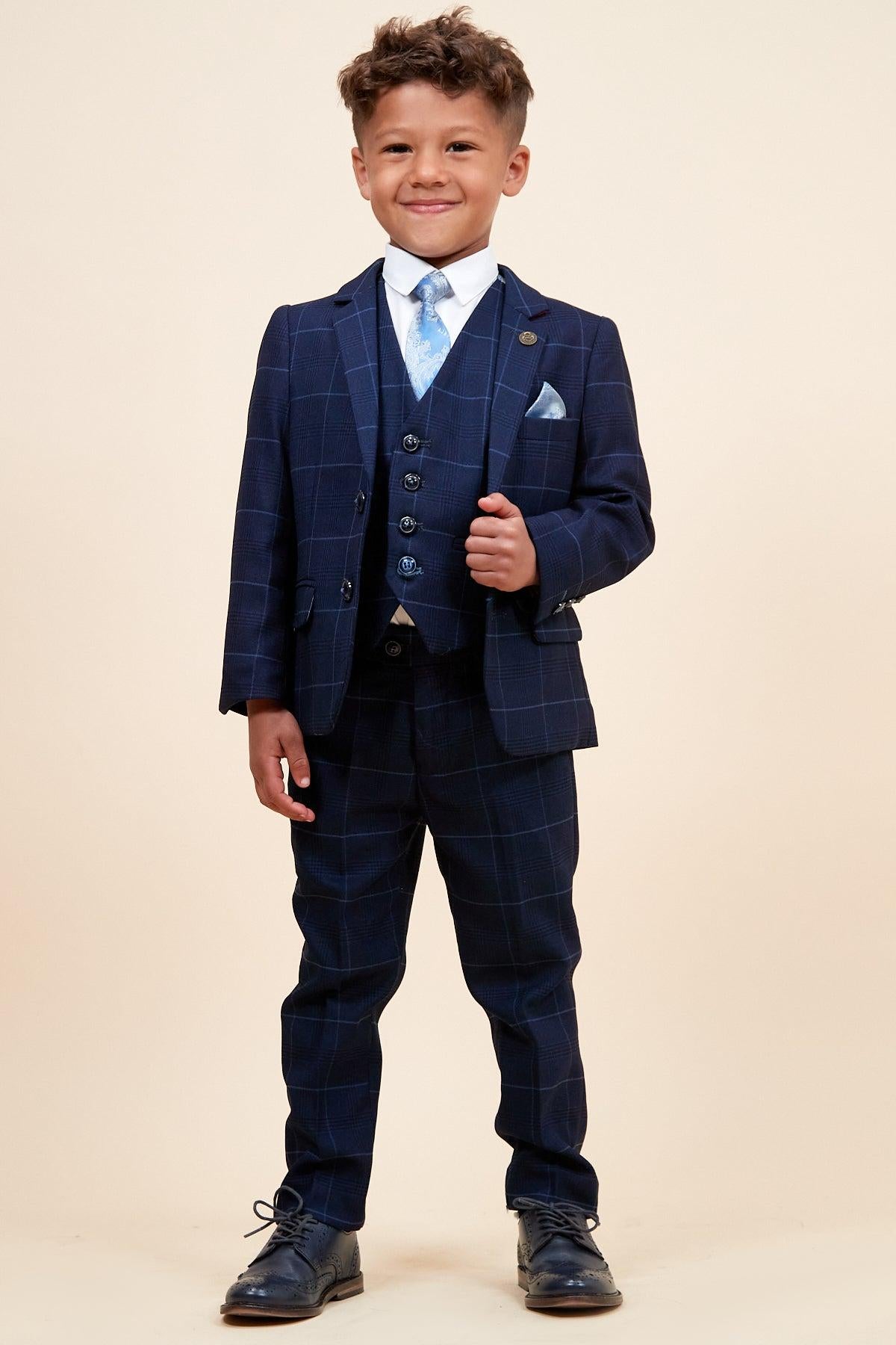 A young boy smiles in the Marc Darcy EDINSON - Children's Navy Sky Check Print Three Piece Suit, complete with matching vest, light blue tie, white shirt, and pocket square, perfect as a kids wedding suit against a plain background.