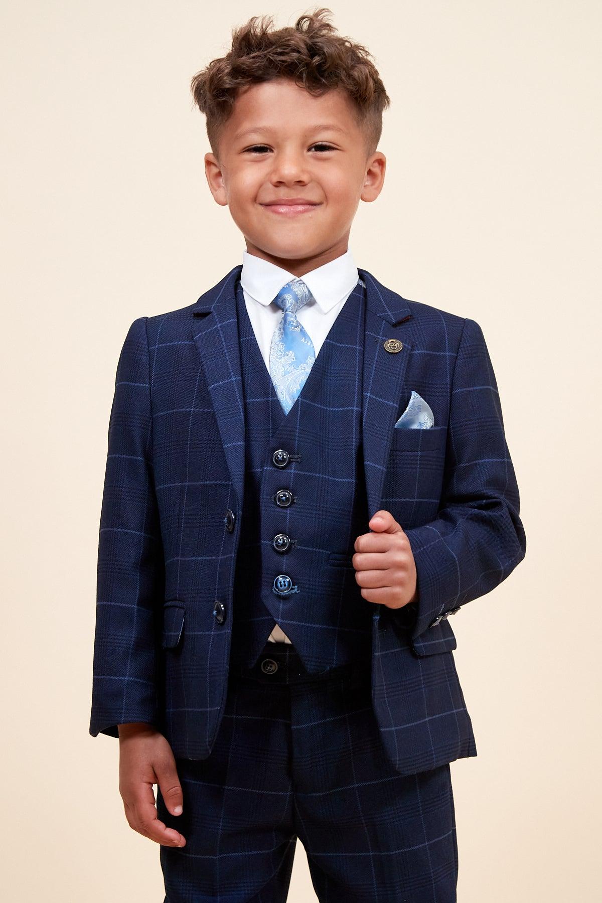 A young boy beams in the Marc Darcy EDINSON—Children’s Navy Sky Check Print Three Piece Suit, a stylish choice for kids featuring a plaid vest, white shirt, and light blue paisley tie—ideal as a children’s wedding suit.