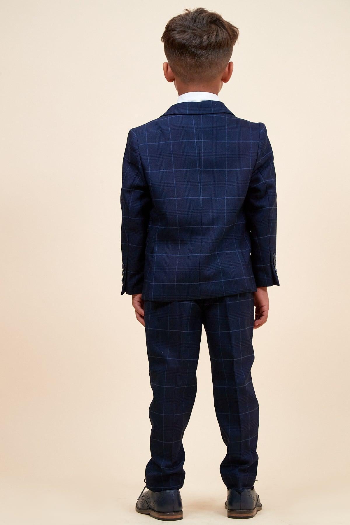 A young boy faces away from the camera, wearing the Marc Darcy EDINSON Children's Navy Sky Check Print Three Piece Suit with matching pants and black shoes, set against a plain beige background.