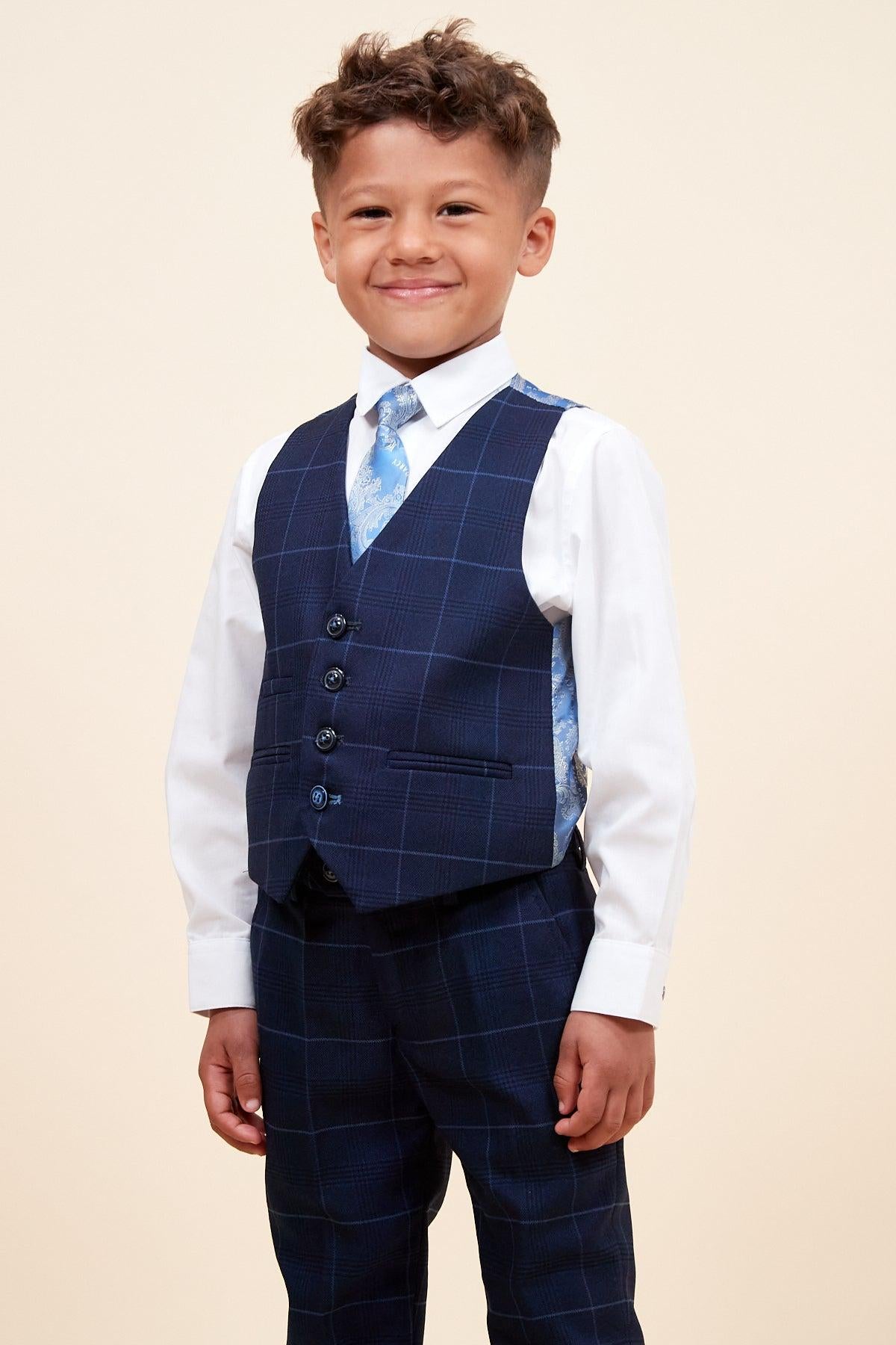 A young boy smiles, dressed in the Marc Darcy EDINSON—Children's Navy Sky Check Print Three Piece Suit—featuring a matching vest, white shirt, and light blue tie, perfect as a kids’ wedding suit against a plain beige background.