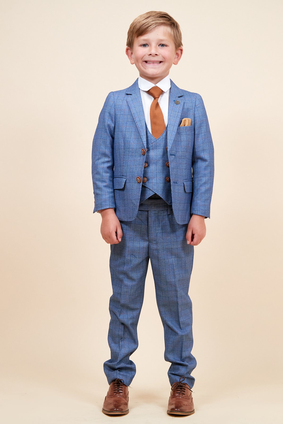 A young boy smiles against a plain background, wearing the marcdarcy GEORGE Children's Light Blue Check Three Piece Suit, styled with a double-breasted waistcoat, white shirt, brown tie, pocket square, and brown dress shoes.