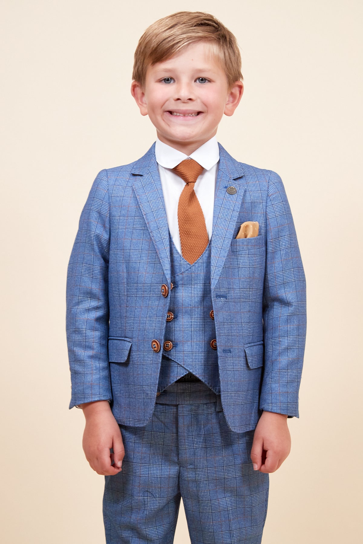 A young boy smiles in front of a beige background, wearing the marcdarcy GEORGE Childrens Light Blue Check Three Piece Suit with double-breasted waistcoat, white shirt, brown tie, and matching pocket square.
