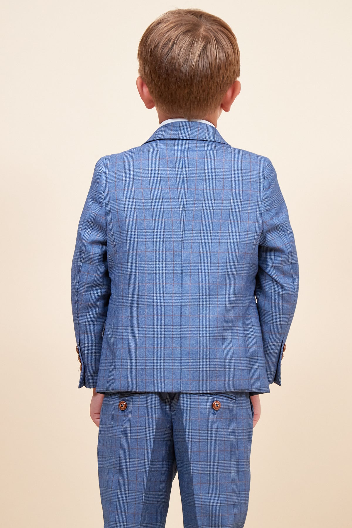 A young boy with light brown hair faces away from the camera, wearing the marcdarcy GEORGE Childrens Light Blue Check Three Piece Suit. The background is plain beige.