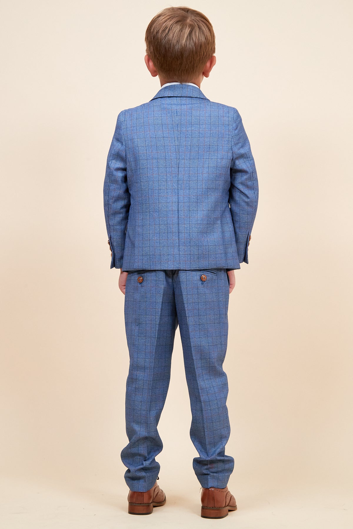 A young boy faces away from the camera, dressed in the marcdarcy GEORGE childrens light blue check three-piece suit with matching trousers and brown shoes, set against a plain beige background.