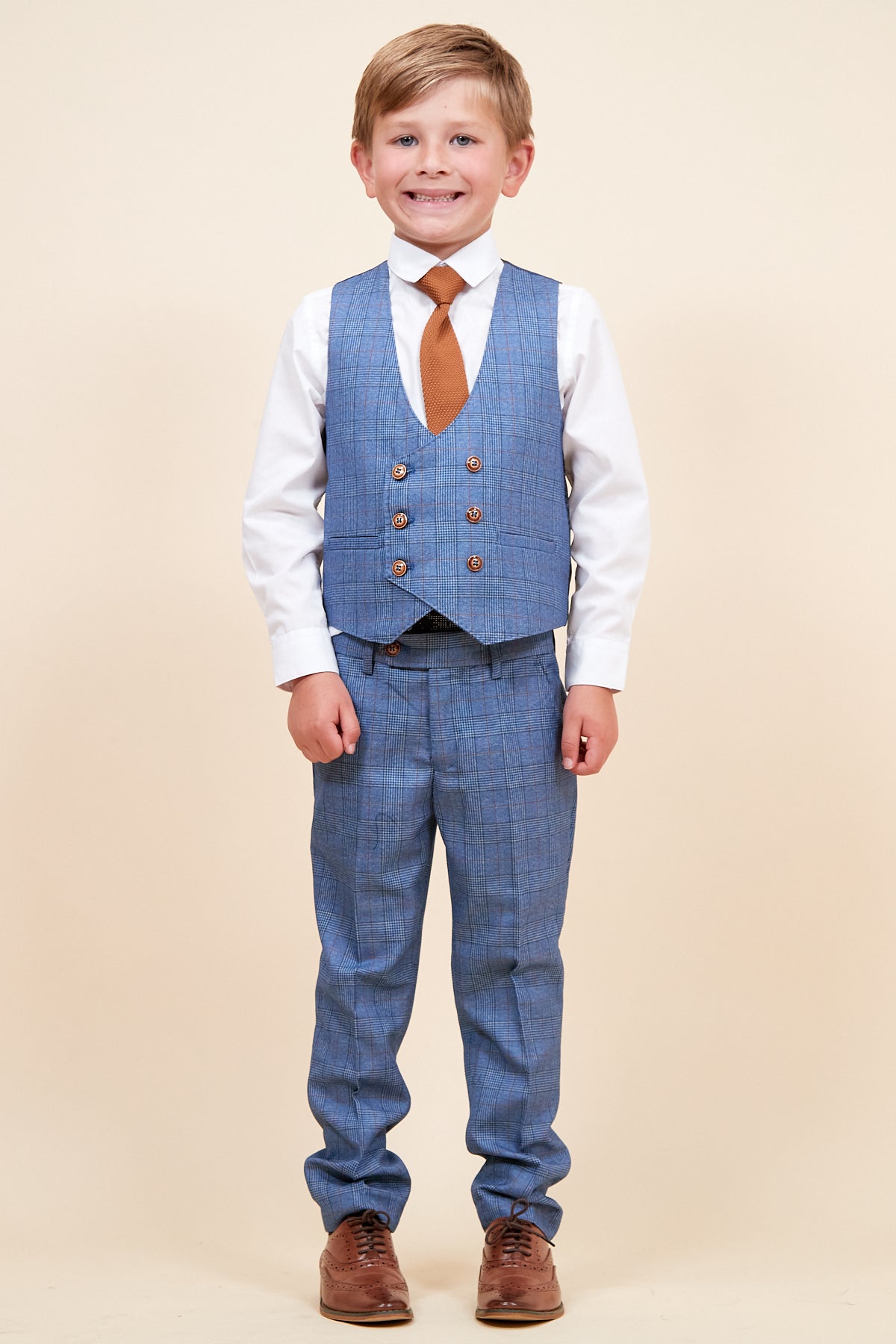 A young boy smiles and poses in the marcdarcy GEORGE children's light blue check three piece suit, paired with a white shirt and burnt orange tie.