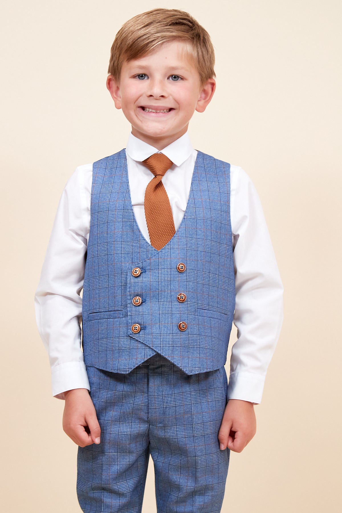 A young boy smiles wearing the marcdarcy GEORGE Childrens Light Blue Check Three Piece Suit, which includes a waistcoat and matching trousers, a white shirt, and brown tie, standing against a plain beige background.
