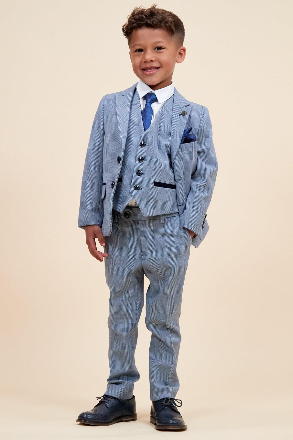 A young boy smiles in the Marc Darcy BROMLEY Children's Sky Blue Check Print Three Piece Suit, featuring a vest, white shirt, blue tie, and dark dress shoes, standing against a plain beige background.