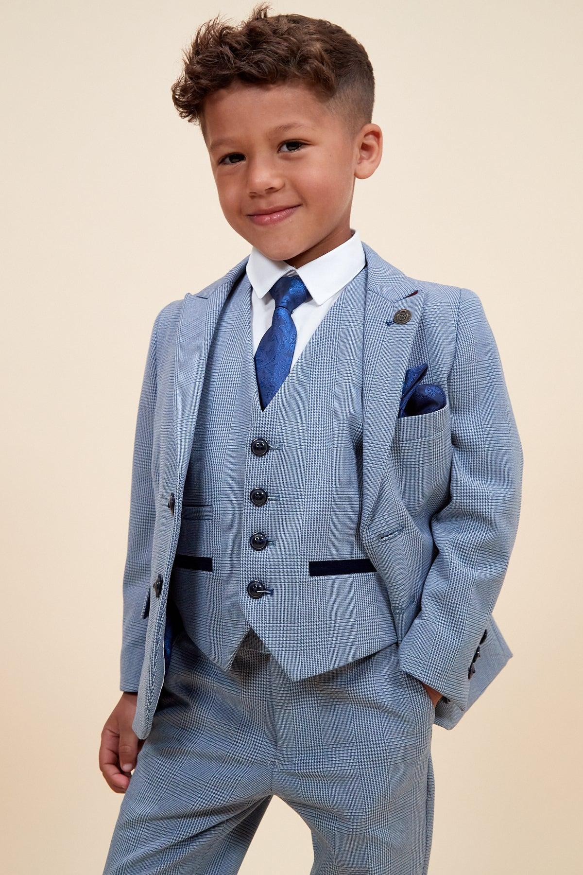 A young boy smiles confidently in the Marc Darcy BROMLEY Children's Sky Blue Check Print Three Piece Suit, featuring a matching vest, navy tie, and white dress shirt against a neutral background.