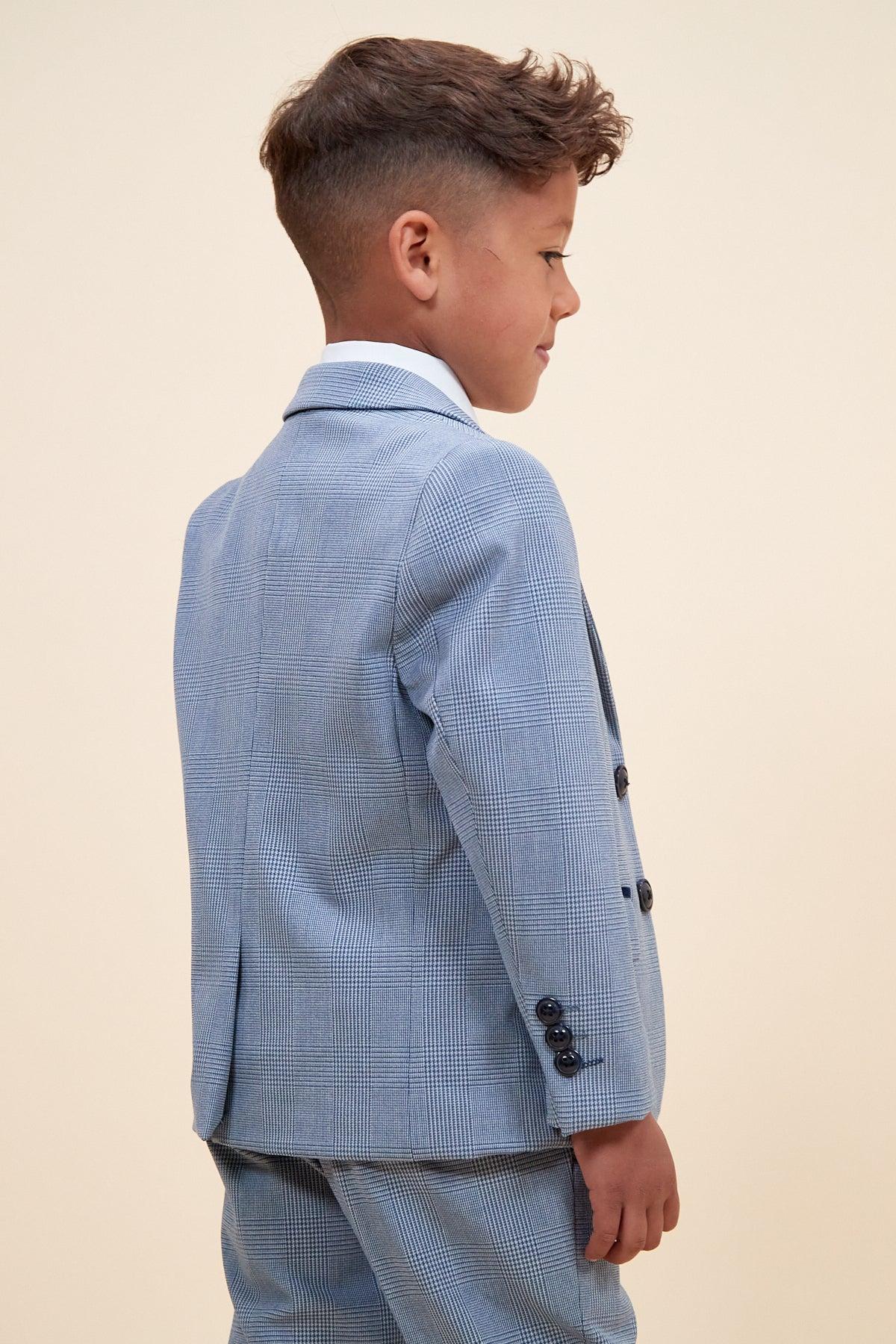 A young boy with short brown hair, dressed in the Marc Darcy BROMLEY Children's Sky Blue Check Print Three Piece Suit and a white shirt, stands sideways and smiles against a plain beige background.
