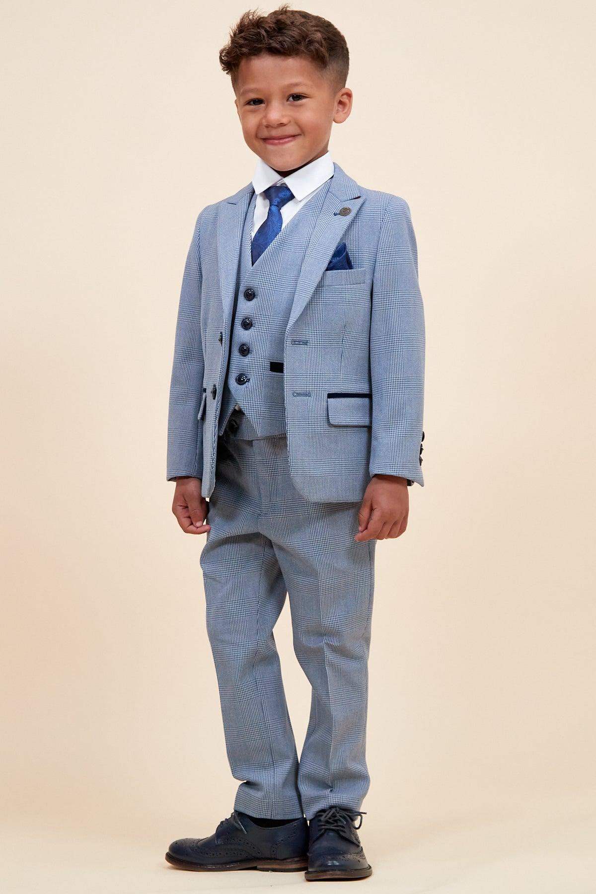 A young boy smiles in the Marc Darcy BROMLEY Children's Sky Blue Check Print Three Piece Suit, styled with a white shirt, blue tie, and blue shoes, standing against a plain beige background.