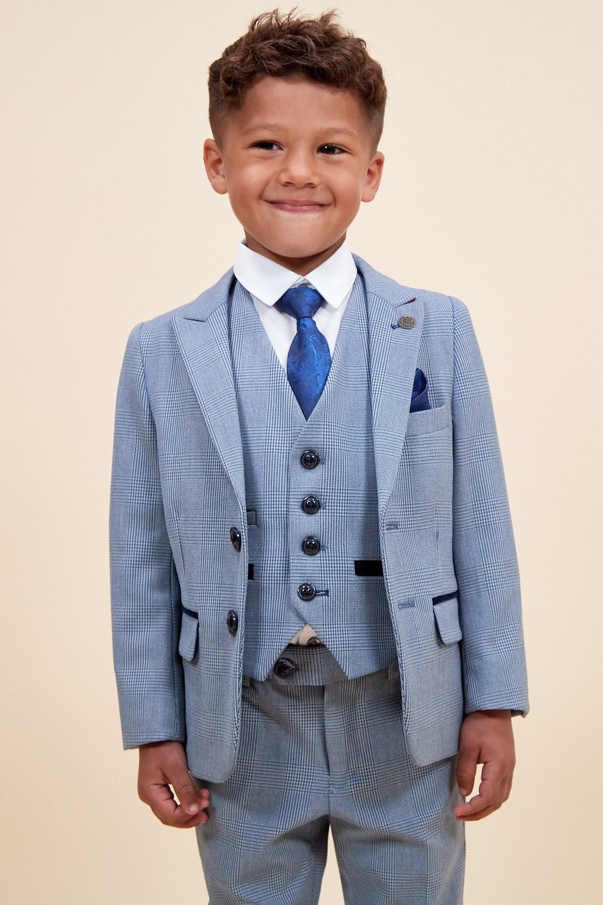 A young boy smiles in the Marc Darcy BROMLEY Children’s Sky Blue Check Print Three Piece Suit with matching vest, white shirt, blue patterned tie, and pocket square, standing against a plain beige background.