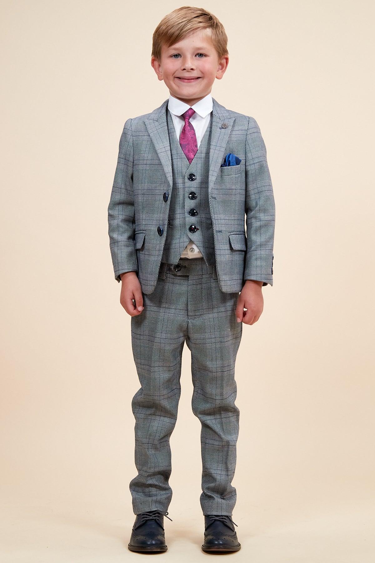 A young boy smiles in the marcdarcy JERRY Childrens Grey Check Three Piece Suit with matching vest, white shirt, purple tie, blue pocket square, and black shoes. The adjustable waistband offers a comfortable fit against a light background.