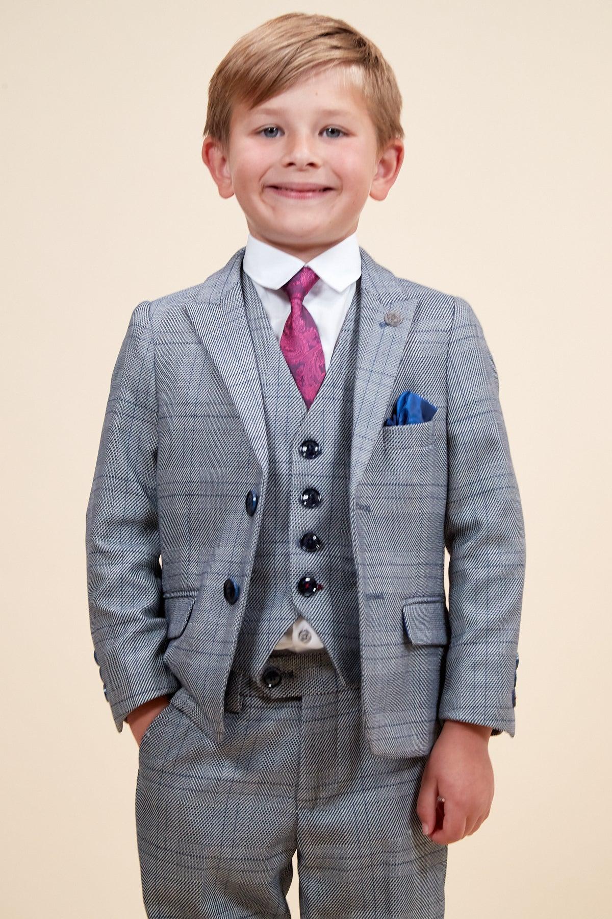 A young boy smiles against a plain background, wearing the marcdarcy JERRY Children's Grey Check Three Piece Suit with an adjustable waistband, matching vest, white shirt, purple patterned tie, and blue pocket square.