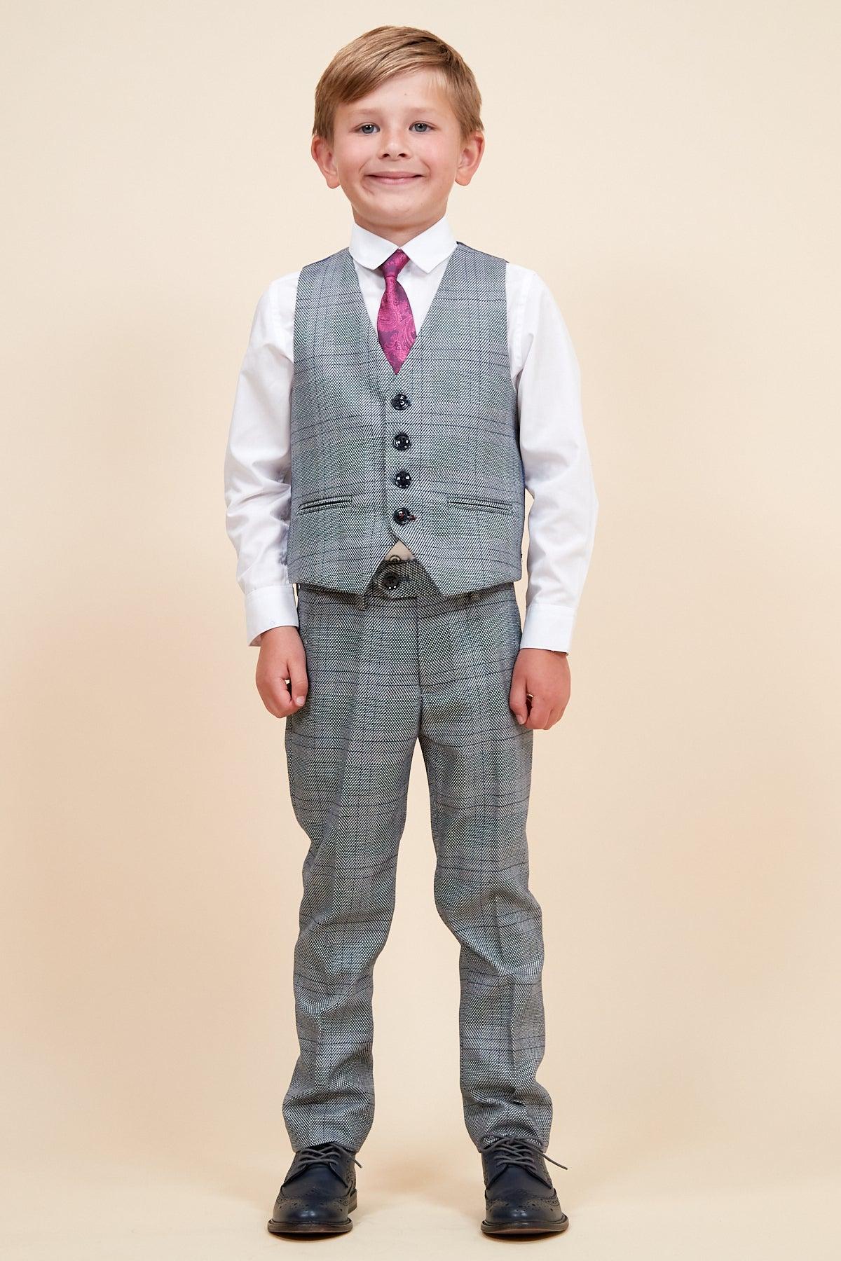 A young boy smiles wearing the marcdarcy JERRY - Childrens Grey Check Three Piece Suit with a matching vest and pants, adjustable waistband, white shirt, pink tie, black shoes, standing against a beige background.