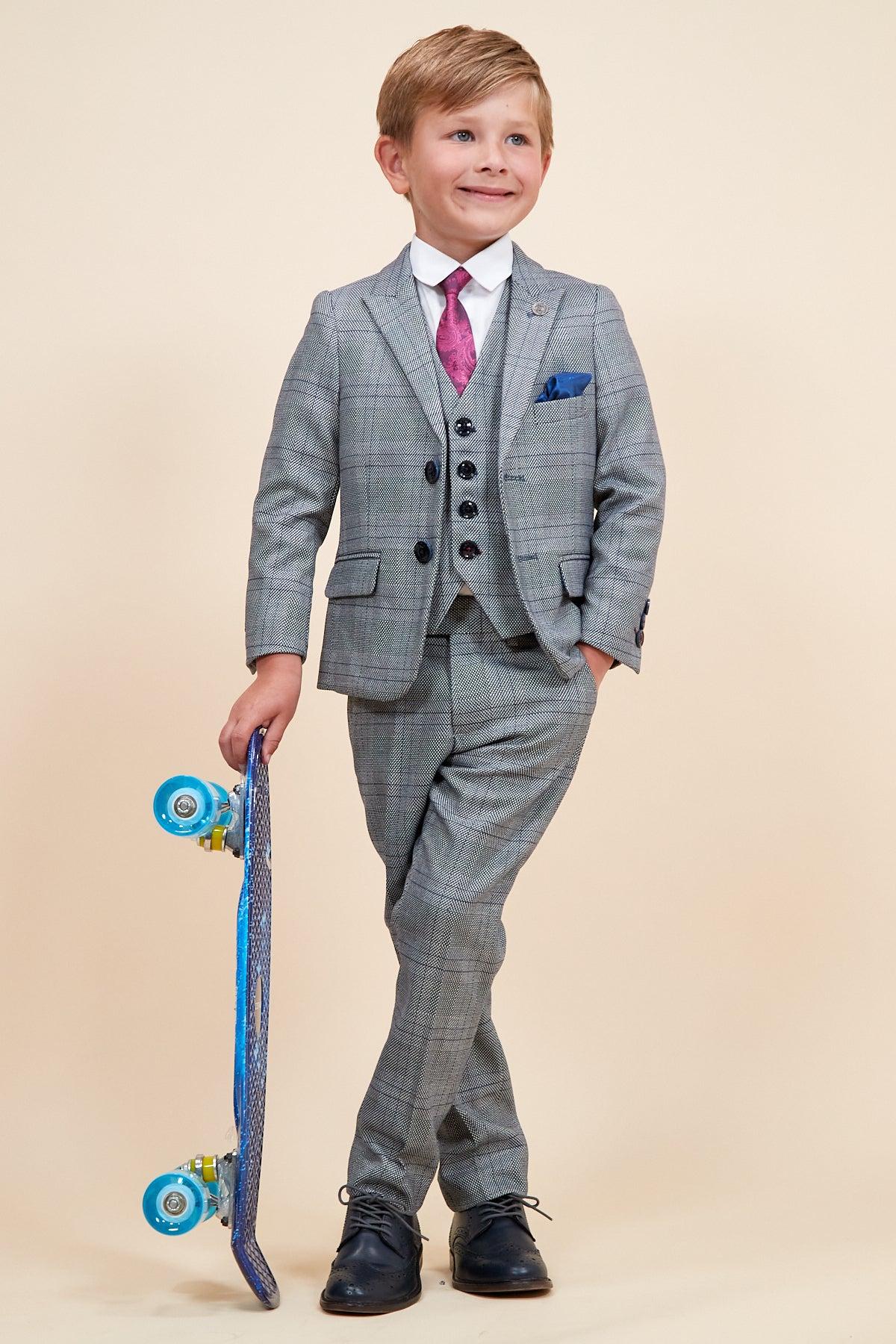 A young boy in the marcdarcy JERRY Children's Grey Check Three Piece Suit, paired with a white shirt and pink tie, stands smiling with a blue skateboard against a plain beige background, one leg crossed over the other.