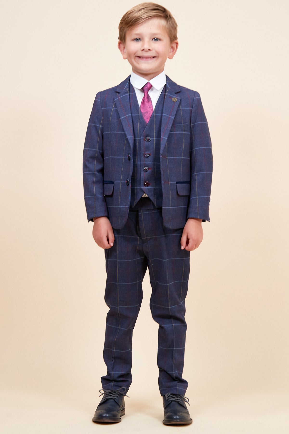 A young boy smiles in the marcdarcy DRAKE Children's Navy Check Three Piece Suit with adjustable waistband, matching vest, white shirt, pink tie, and dark dress shoes against a plain beige background.