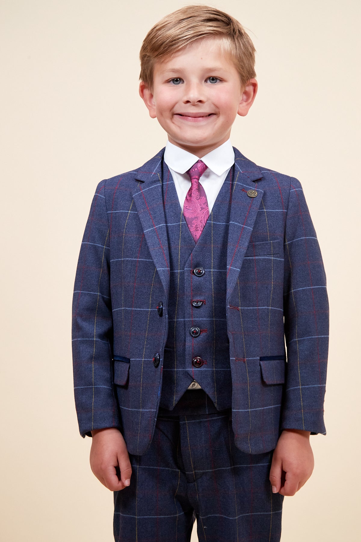 A young boy with blonde hair smiles in the marcdarcy DRAKE Childrens Navy Check Three Piece Suit, featuring a vest, white shirt, and pink patterned tie, set against a plain light background.
