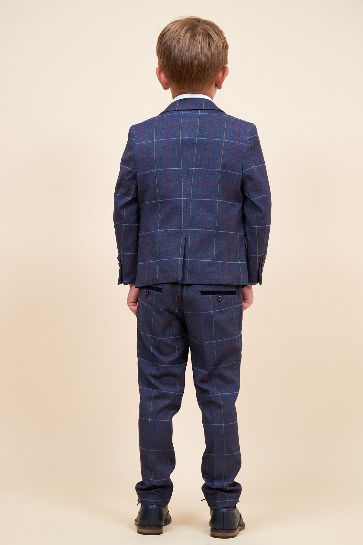 A young boy faces away from the camera, wearing the marcdarcy DRAKE - Children's Navy Check Three Piece Suit with adjustable waistband and matching trousers, plus black dress shoes, set against a plain beige background.