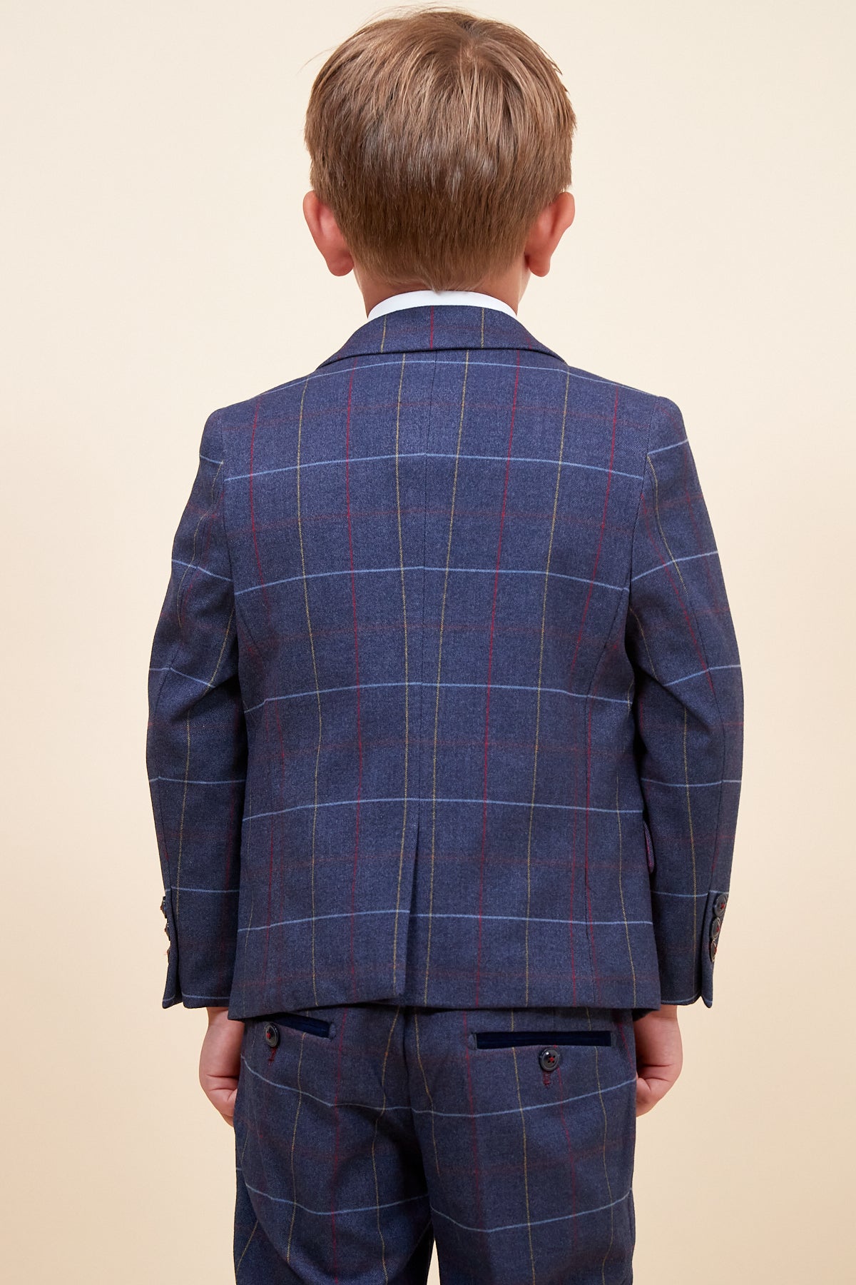A young boy stands facing away in the marcdarcy DRAKE Children’s Navy Check Three Piece Suit, featuring a fitted jacket and matching trousers with red and yellow lines, creating a smart pageboy look against a light background.