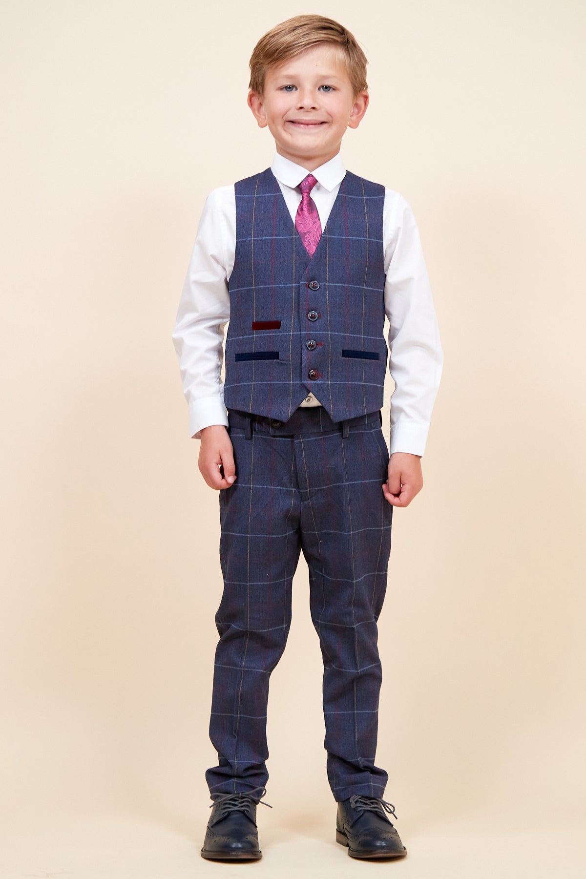 A young boy smiles in the marcdarcy DRAKE Childrens Navy Check Three Piece Suit, featuring a navy check jacket and matching vest, white shirt, pink patterned tie, and black dress shoes for a sharp pageboy look against a beige background.