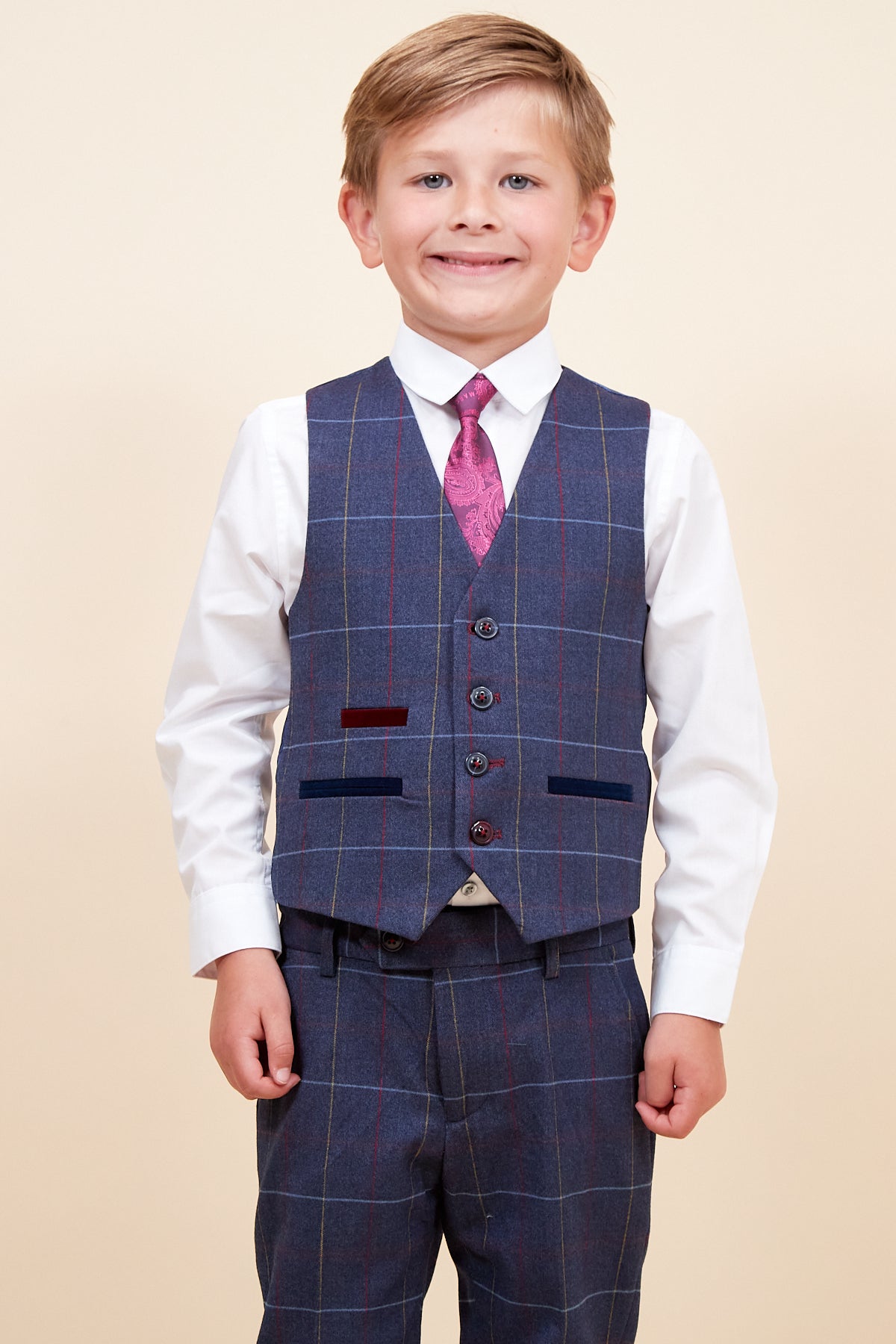 A young blonde boy smiles in the marcdarcy DRAKE Childrens Navy Check Three Piece Suit—featuring a white shirt, pink patterned tie, and matching vest and trousers—posing against a plain beige background.